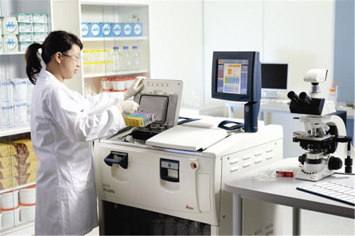 A tissue processor being loaded with a basket of cassettes containing tissue specimens for processing. Details of the processing steps and the schedule are shown on the screen of the processor.